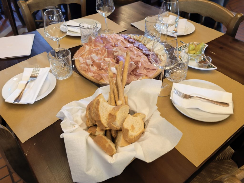 Verpflegung auf einem regionalen Weingut. Hochwertiges Weißbrot und Brot Sticks in einem Korb umgeben von Gedeck. Dahinter eine Platte mit Italienischem Schinken, Salami und Käse.